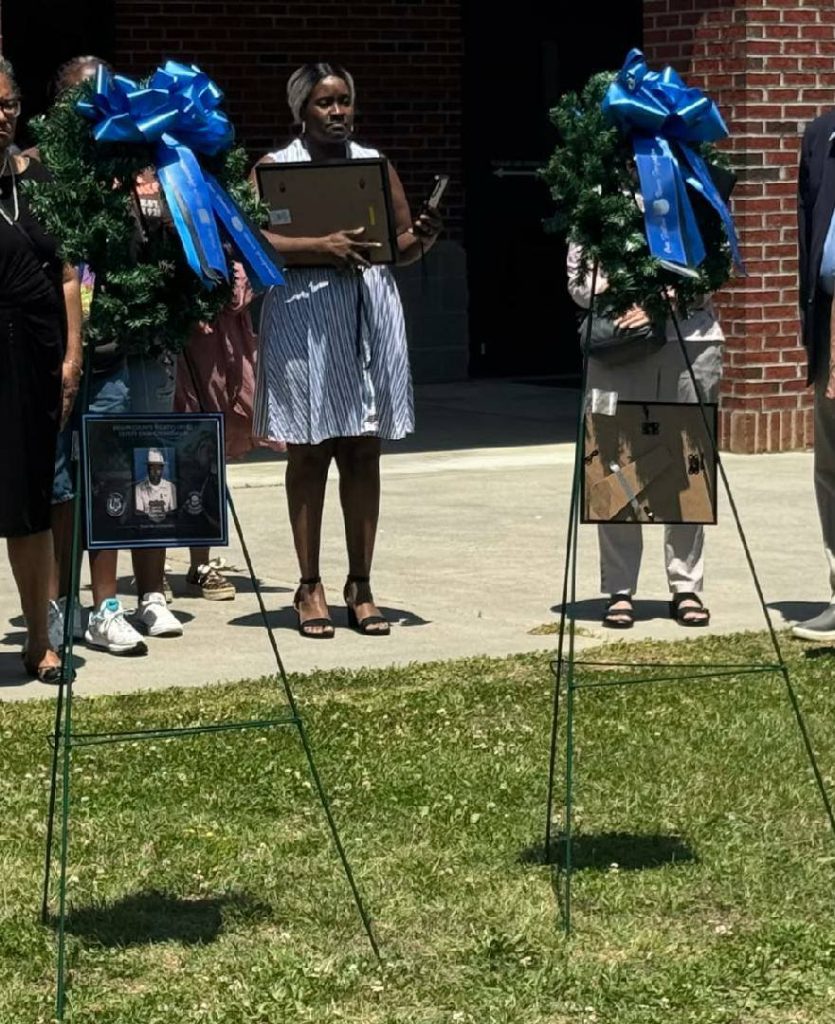 People stand outdoors near two memorial wreaths with large blue bows and framed photos, during what appears to be a commemorative or memorial event.