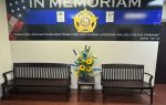 Memorial wall with police badge, American flag, two benches, a flower arrangement, and Bible verse honoring fallen officers.
