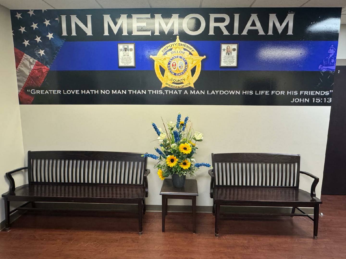 Memorial wall with police badge, American flag, two benches, a flower arrangement, and Bible verse honoring fallen officers.