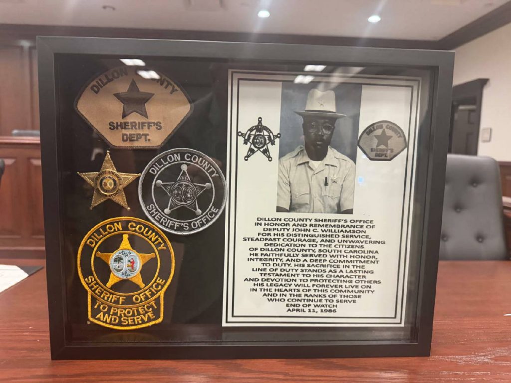 Framed display with sheriff badges, a black and white photograph of an officer, and a tribute text honoring Deputy John C. Williamson from Dillon County Sheriff’s Office.