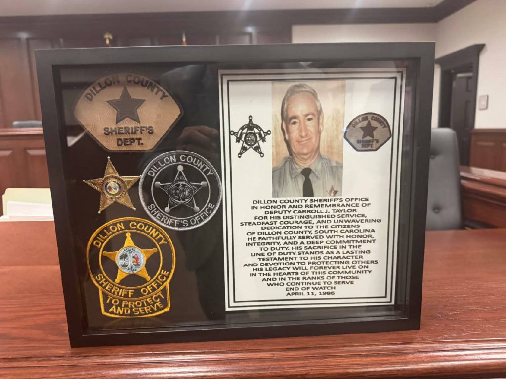A framed display honors Deputy Carroll J. Taylor with a photo, badges, and a tribute from Dillon County Sheriff's Office, placed on a wooden desk in a courtroom.