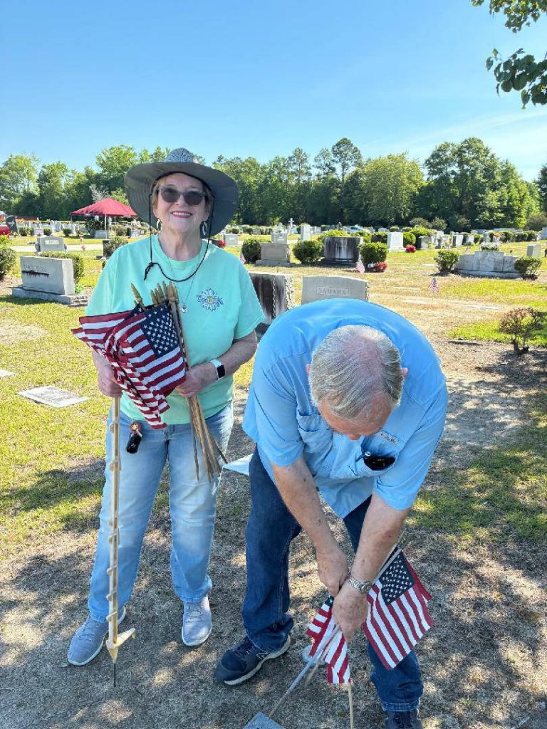 Two people placing American flags on graves in a cemetery on a sunny day, honoring veterans or Memorial Day.