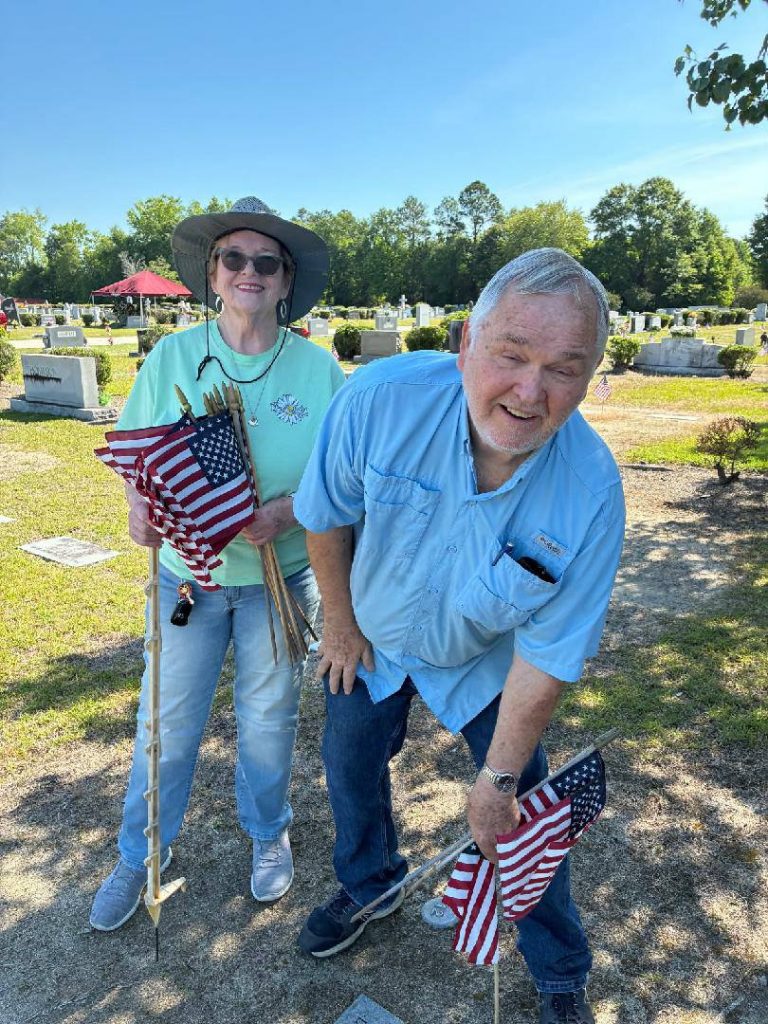 Two older adults placing American flags on graves in a cemetery on a sunny day, honoring veterans.
