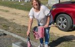 Smiling woman places American flag at a gravestone in a cemetery, holding several more flags, with a red car parked nearby.
