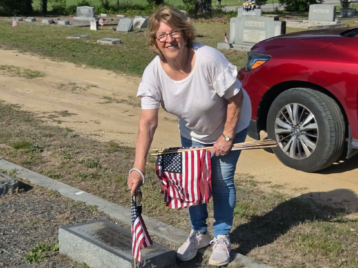Smiling woman places American flag at a gravestone in a cemetery, holding several more flags, with a red car parked nearby.