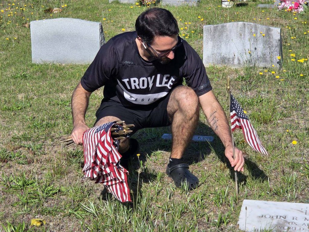 A man kneels at a grave, placing small American flags on a grassy cemetery plot under bright sunlight.