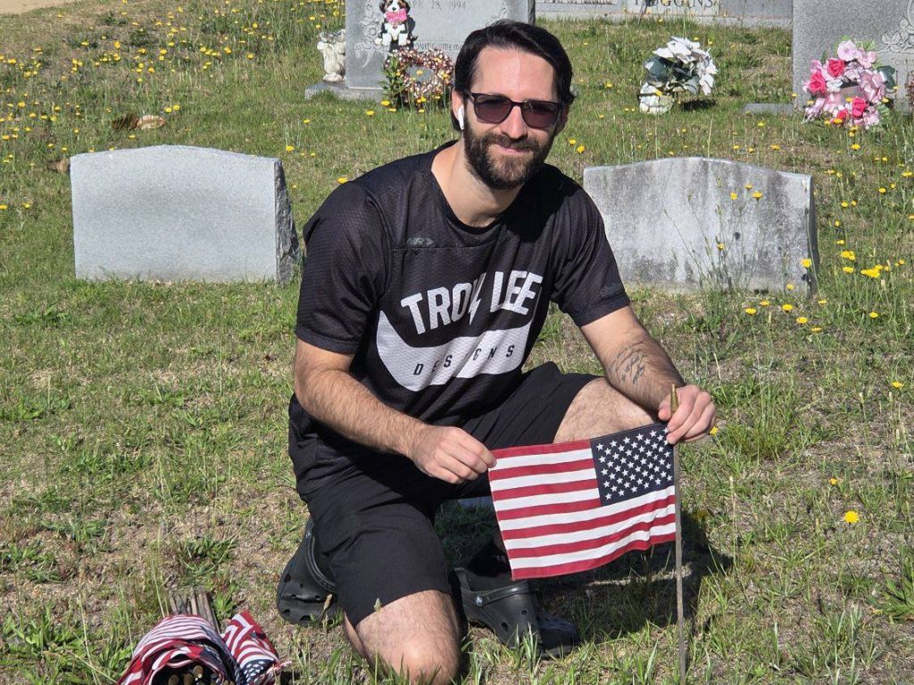 Man kneeling in a cemetery, holding an American flag, wearing sunglasses and a black shirt. Gravestones and flowers are visible in the background on a sunny day.