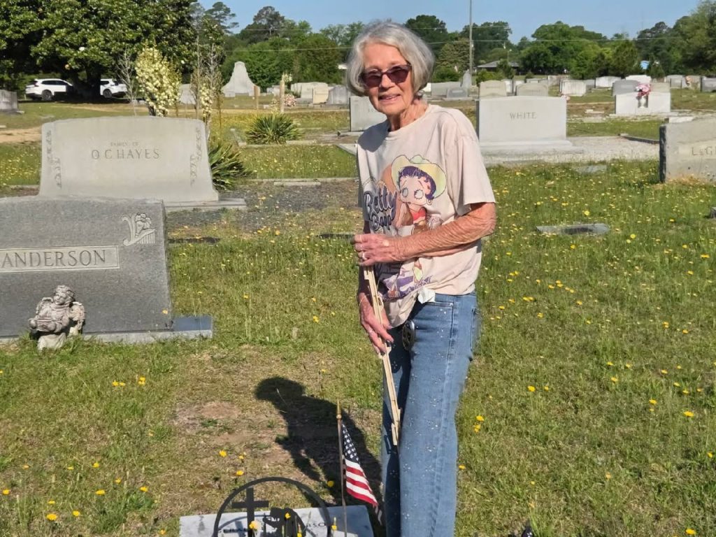 Elderly woman with sunglasses and a Betty Boop shirt stands in a grassy cemetery near a gravestone and an American flag on a sunny day.