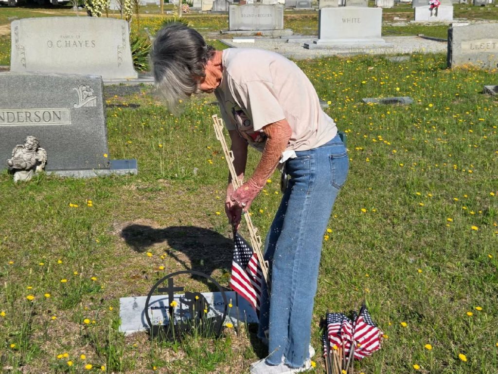 elderly woman places American flags at a grave in a cemetery, surrounded by tombstones and yellow wildflowers on a sunny day.