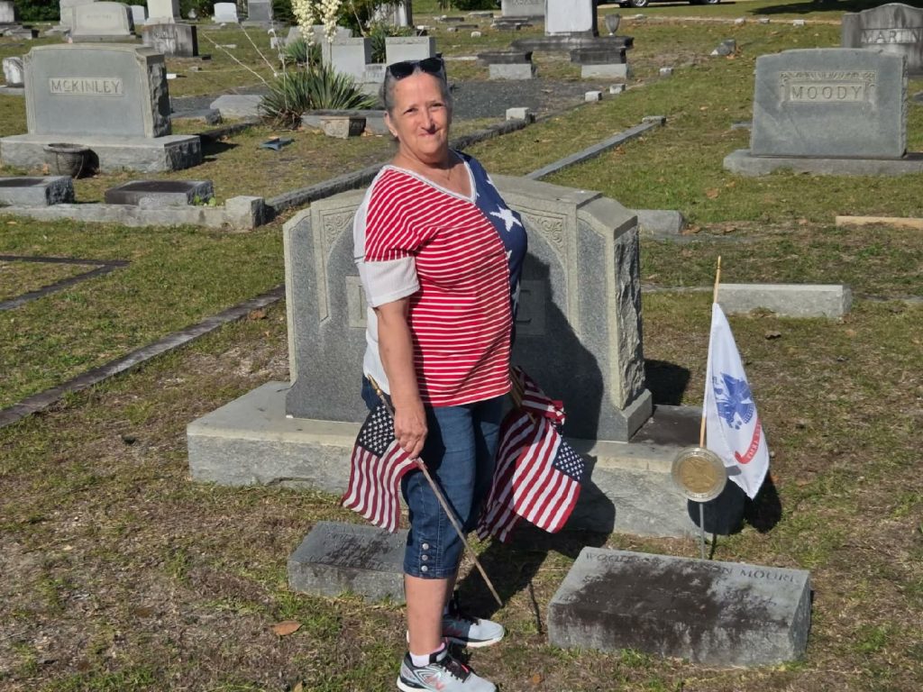 Woman in a red, white, and blue shirt stands in a cemetery holding American flags near a gravestone with a military flag and medallion.
