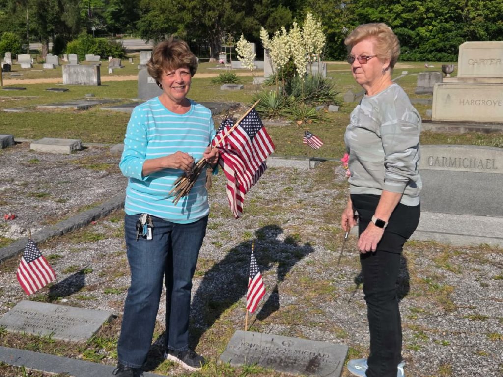 Two women place American flags on graves in a cemetery on a sunny day, honoring veterans with headstones and greenery visible in the background.