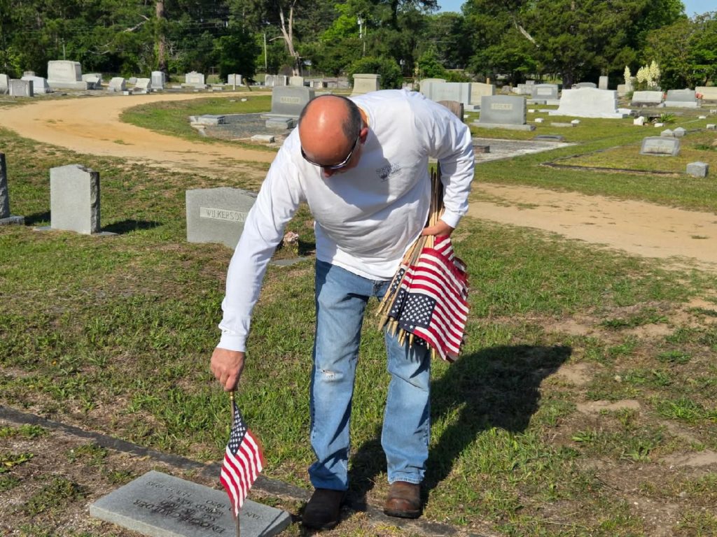 A man places an American flag at a gravesite in a cemetery, holding several more flags in his arm on a sunny day.