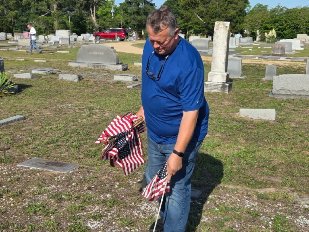 Man in blue shirt placing American flags on graves in a cemetery, with tombstones and a red car visible in the background.