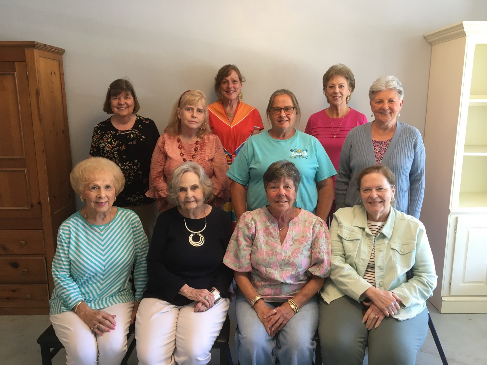 A group of eleven women posing indoors, sitting and standing in three rows, smiling at the camera with neutral background and wooden furniture.