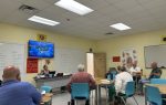 Several adults sit in a classroom, facing a teacher at a desk, with anatomy posters and notes on the whiteboard.