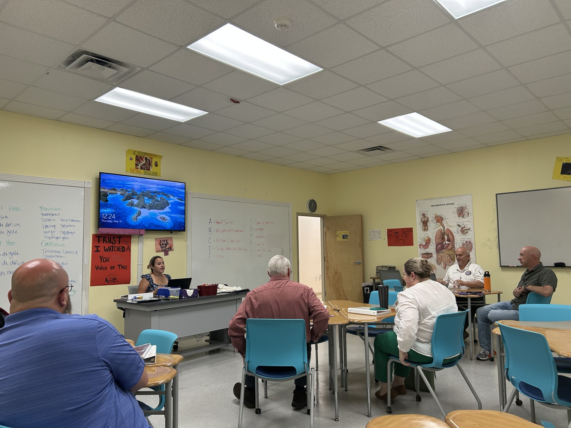 Several adults sit in a classroom, facing a teacher at a desk, with anatomy posters and notes on the whiteboard.