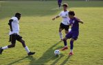 Three soccer players compete for the ball on a grassy field, with one player in purple about to kick while two players in white defend.