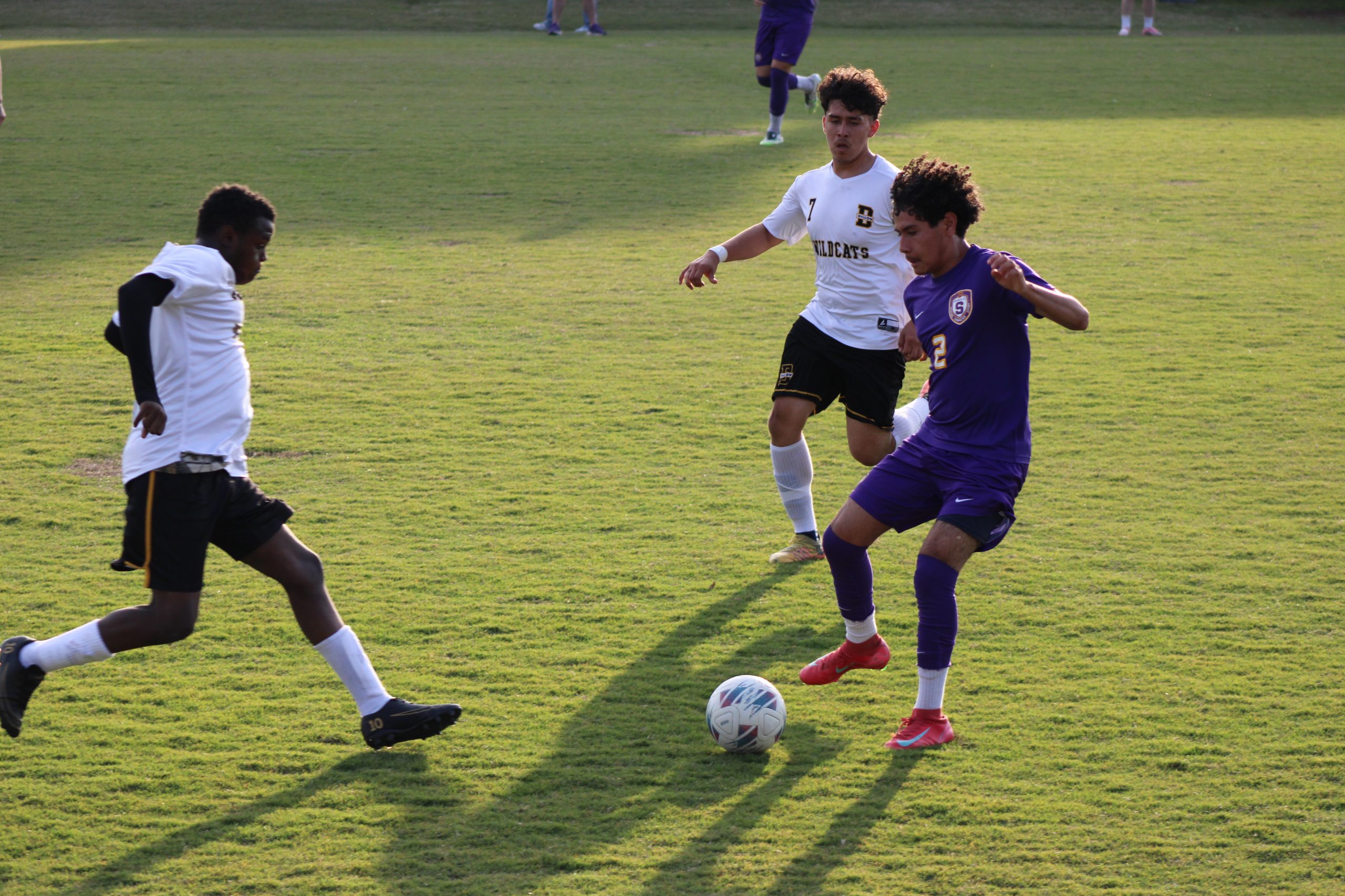 Three soccer players compete for the ball on a grassy field, with one player in purple about to kick while two players in white defend.