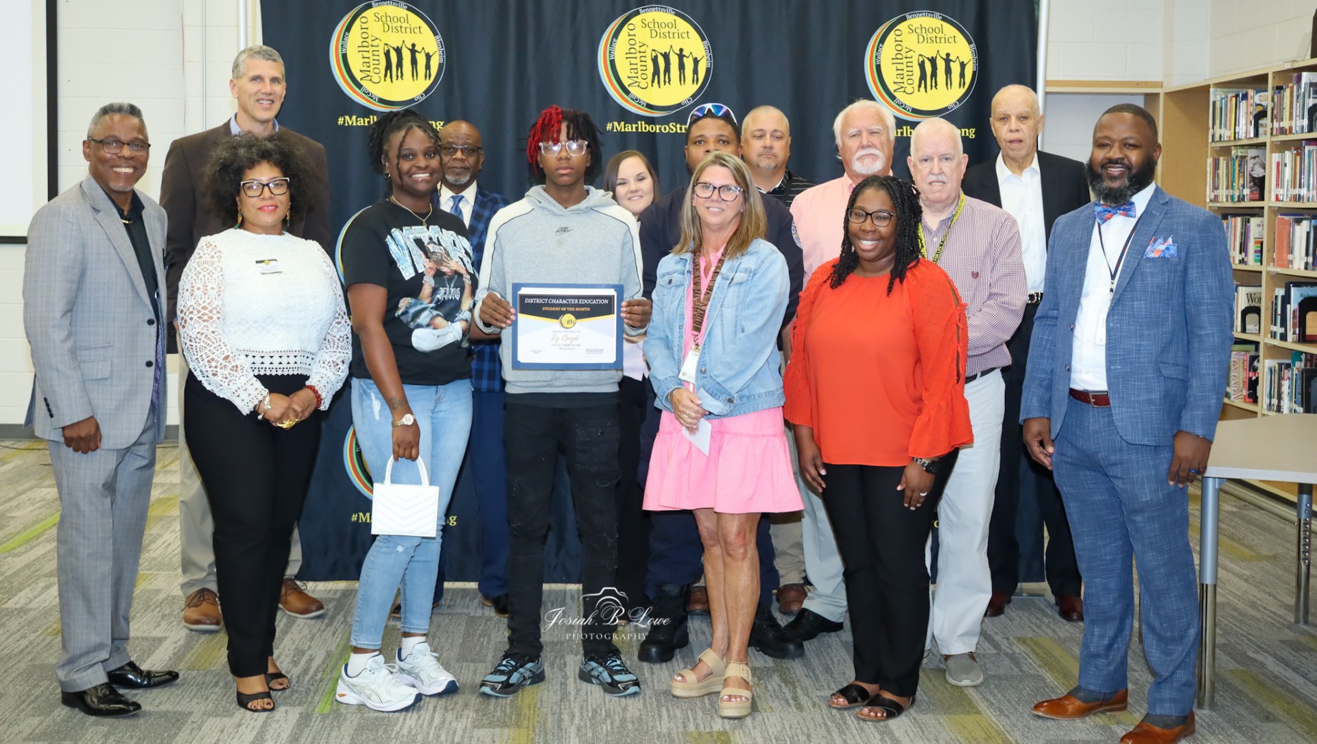 A group of people pose indoors with a young student holding a certificate, standing in front of a Marlboro County School District backdrop.
