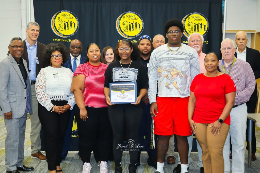 A group of people stands together, with one person holding a certificate, posing for a photo in front of Marlboro School District banners.