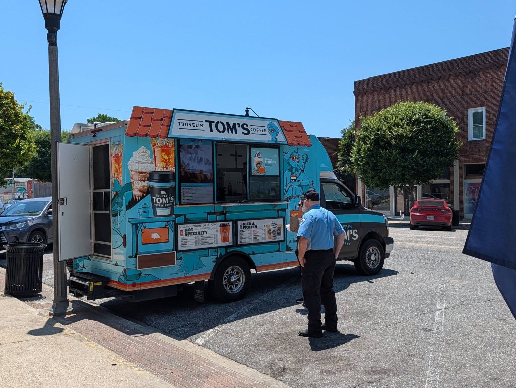 A person stands at a colorful coffee truck labeled "Travelin' Tom's Coffee" on a sunny downtown street with buildings and parked cars in the background.
