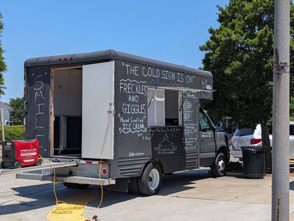 Black food truck with chalk writing advertises “Freckles and Giggles” handcrafted ice cream, parked outdoors on a sunny day near trees and cars.