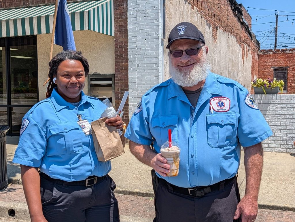 Two paramedics in blue uniforms smiling outdoors, holding drinks and a paper bag, standing on a sunny sidewalk near a brick building.