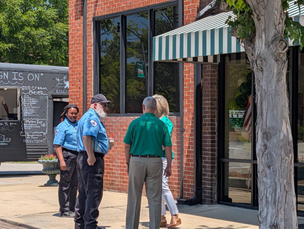 Two uniformed security officers and two civilians stand talking outside a brick building with striped awning, near a food truck and tree on a sunny day.