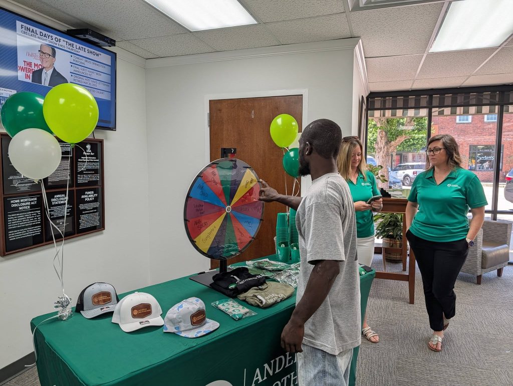 Man spinning prize wheel at a promotional event with balloons, hats, and two women in green shirts watching.