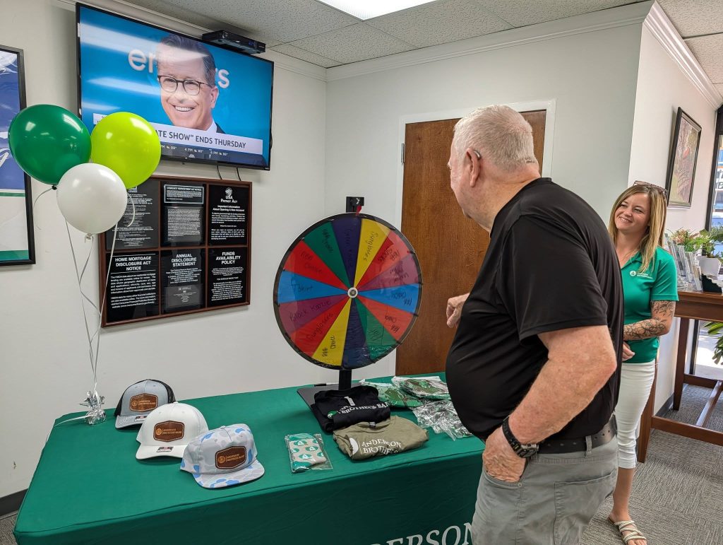 older man spins a colorful prize wheel at a table with hats and gifts, while a smiling woman watches in a decorated office.