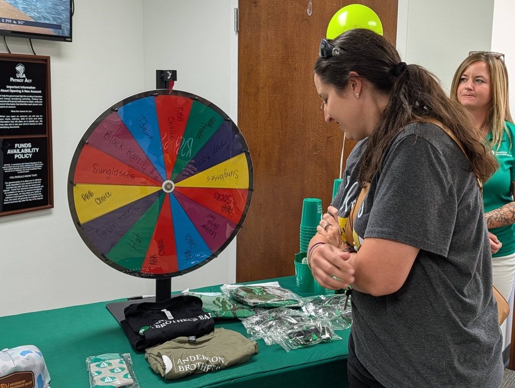 A woman stands near a colorful prize wheel at an event table with shirts, cups, and other promotional items, while another woman observes.