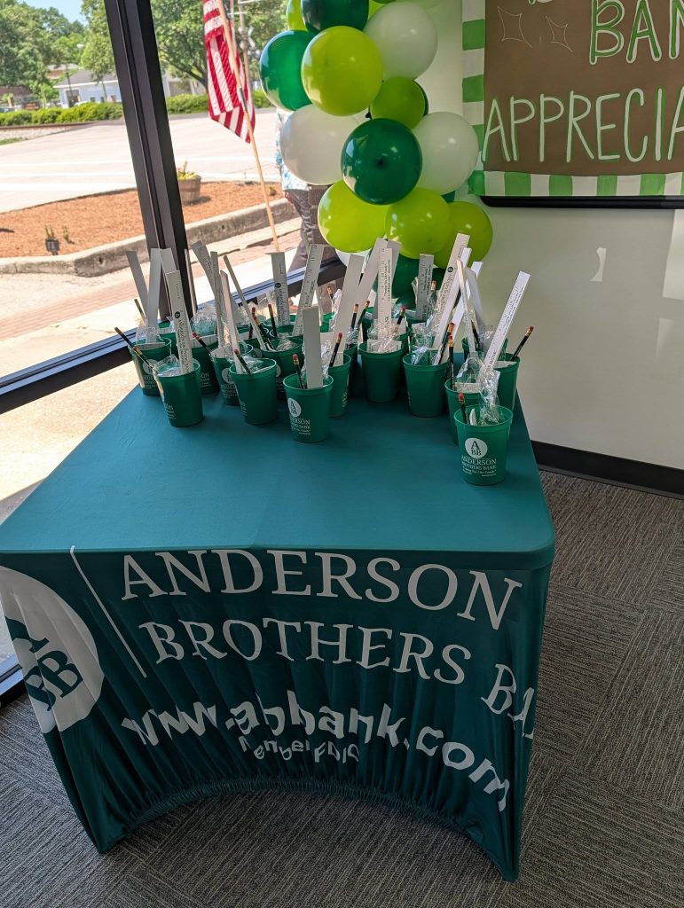 Green table with Anderson Brothers Bank cups filled with pens and rulers, green and white balloons, and an appreciation sign in a sunlit room with an American flag outside.