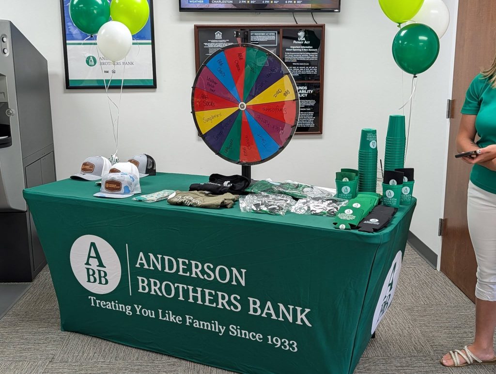 A promotional table for Anderson Brothers Bank with branded items, a prize wheel, green cups, balloons, hats, and a woman standing to the right.