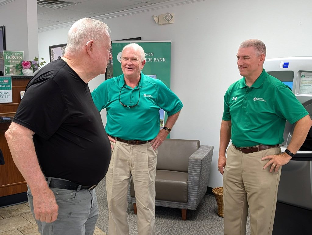 Three men talking inside a bank, two in green shirts and khaki pants, one in black shirt and gray pants, standing near a seating area and ATM.