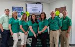 Seven people in green shirts stand together inside an office with balloons and a promotional display in the background.