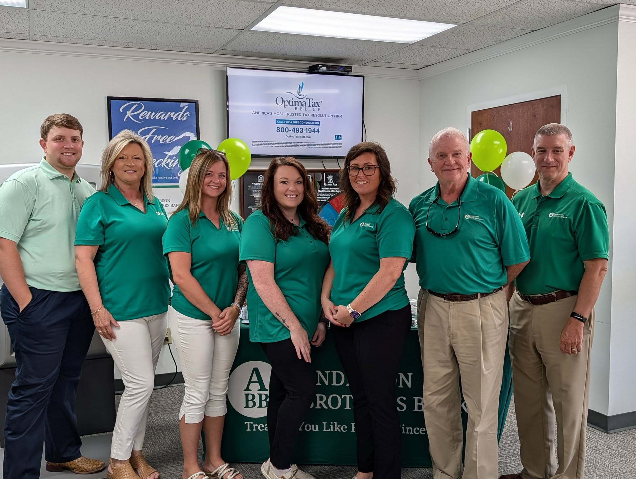Seven people in green shirts stand together inside an office with balloons and a promotional display in the background.