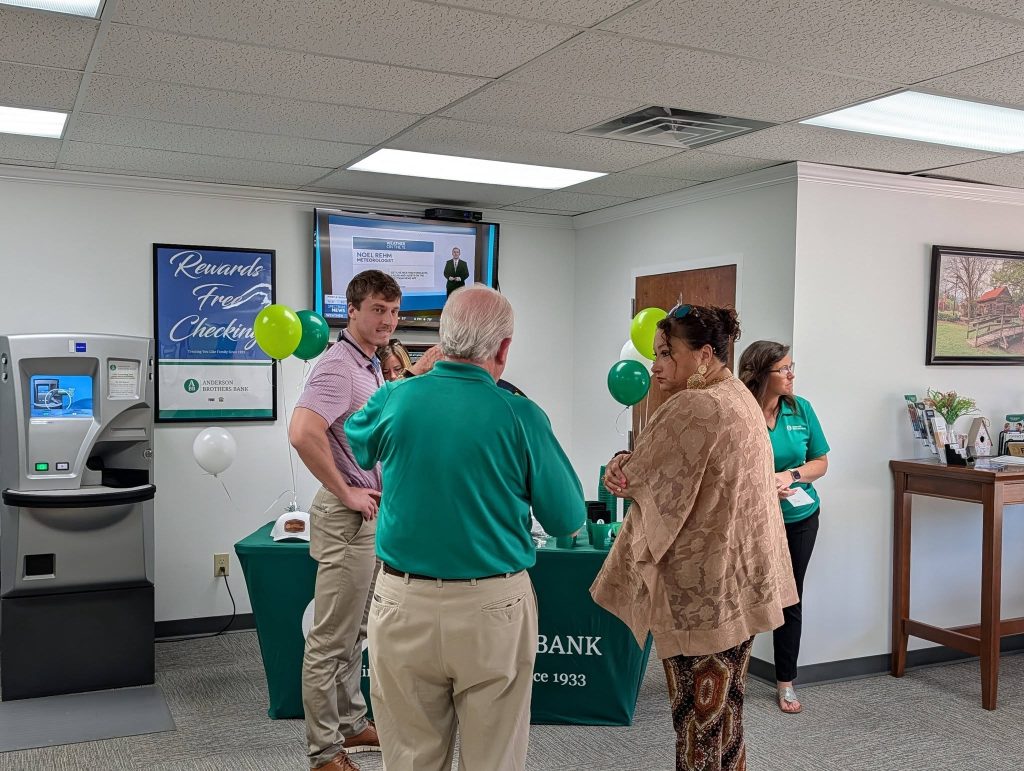 People chatting near a bank booth with balloons and brochures, an ATM on the left, and a TV displaying weather information in the background.