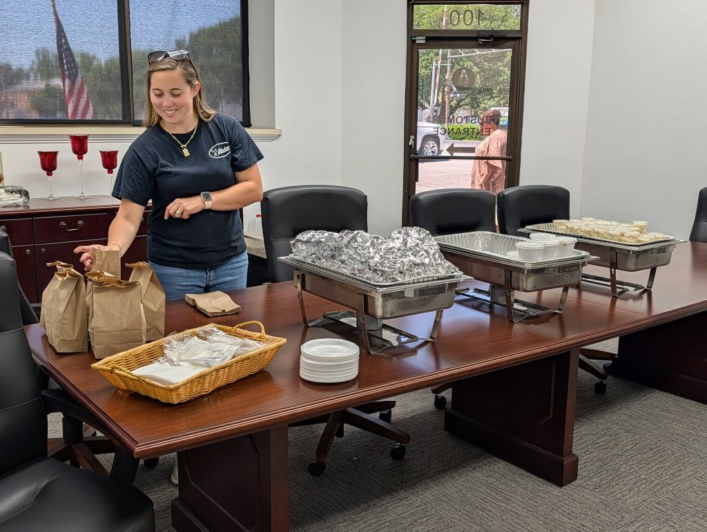 A woman sets up a buffet with covered food trays, brown bags, cups, plates, and utensils in a conference room with large windows.