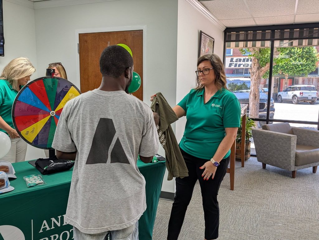 A woman hands a t-shirt to a man standing by a table with a prize wheel at an indoor promotional event.