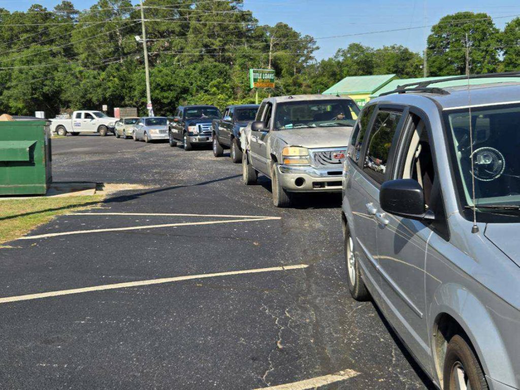Several cars and trucks are lined up in a drive-thru on a sunny day with trees in the background.