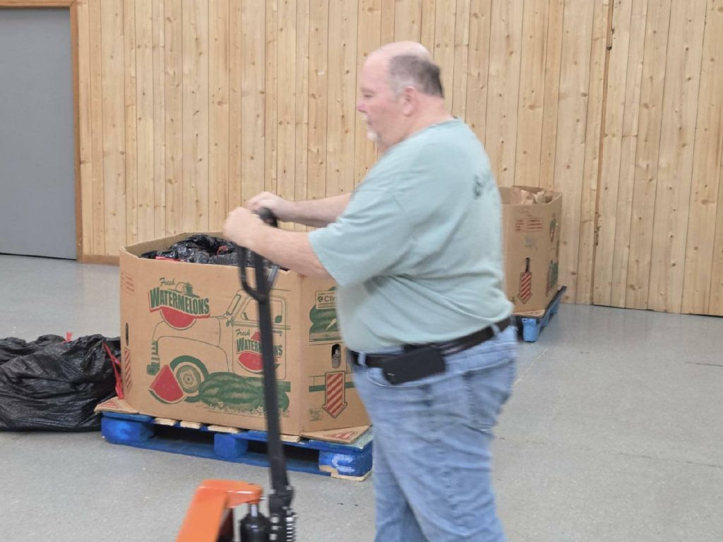 A man moves a pallet jack near large cardboard boxes labeled "Fresh Watermelons" inside a room with wooden walls.