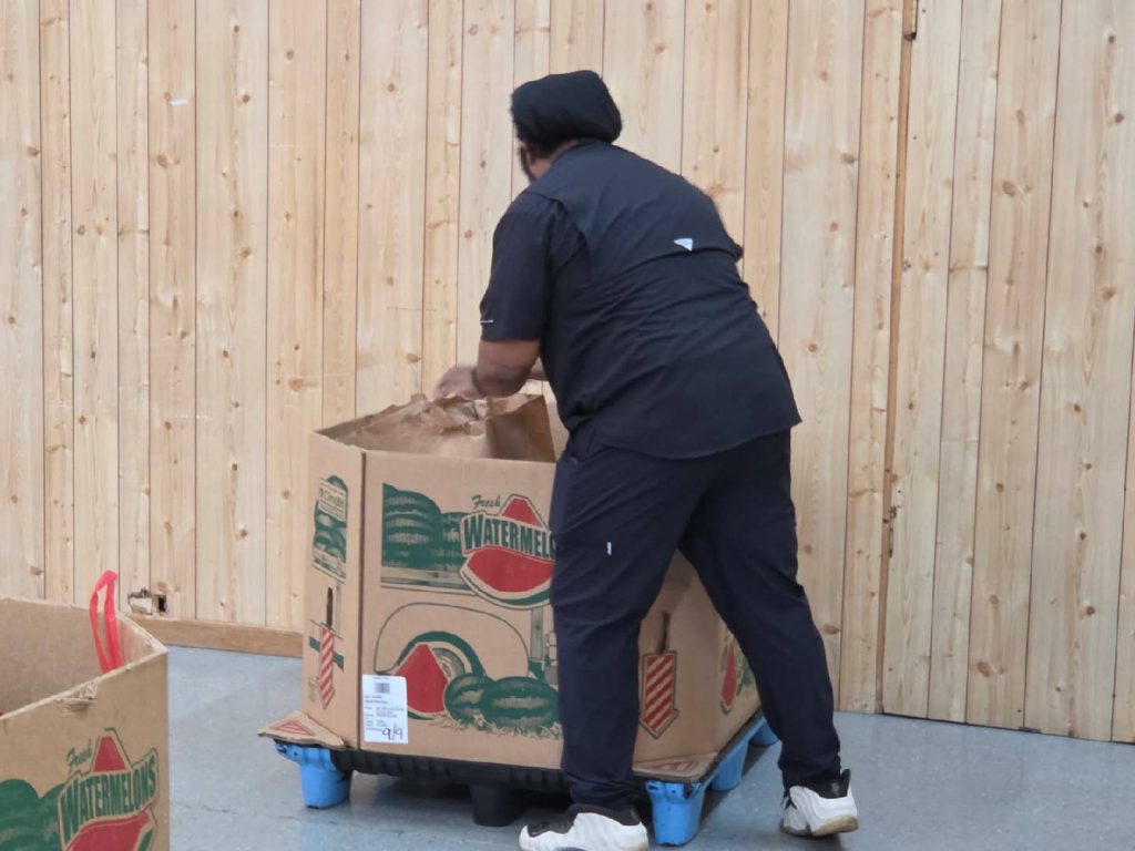 A person in dark clothing handles a large cardboard box labeled “watermelons” placed on a blue pallet, against a wooden wall background.