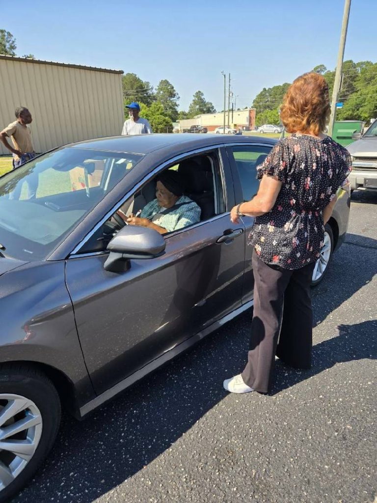 A woman stands by a parked car speaking to a person inside, while two other people stand nearby outdoors on a sunny day.