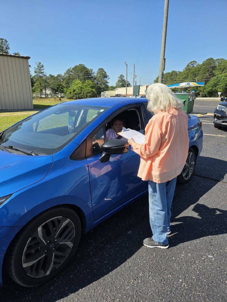 A person stands beside a blue car, talking to the driver and handing over a sheet of paper in a parking lot on a sunny day.