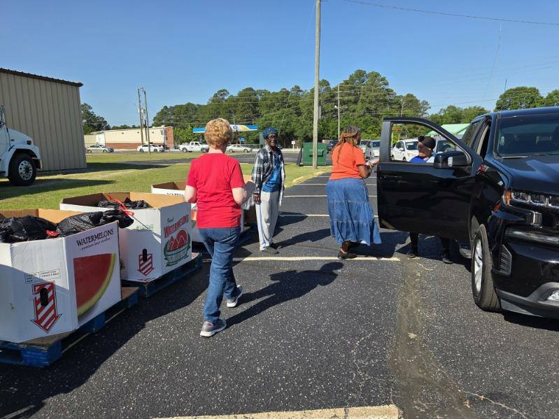 People distribute food from large watermelon boxes in a parking lot, with some placing items into a black SUV on a sunny day.