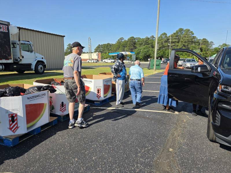 People distributing watermelons from large boxes in a parking lot, with a truck and several cars visible in the background on a sunny day.