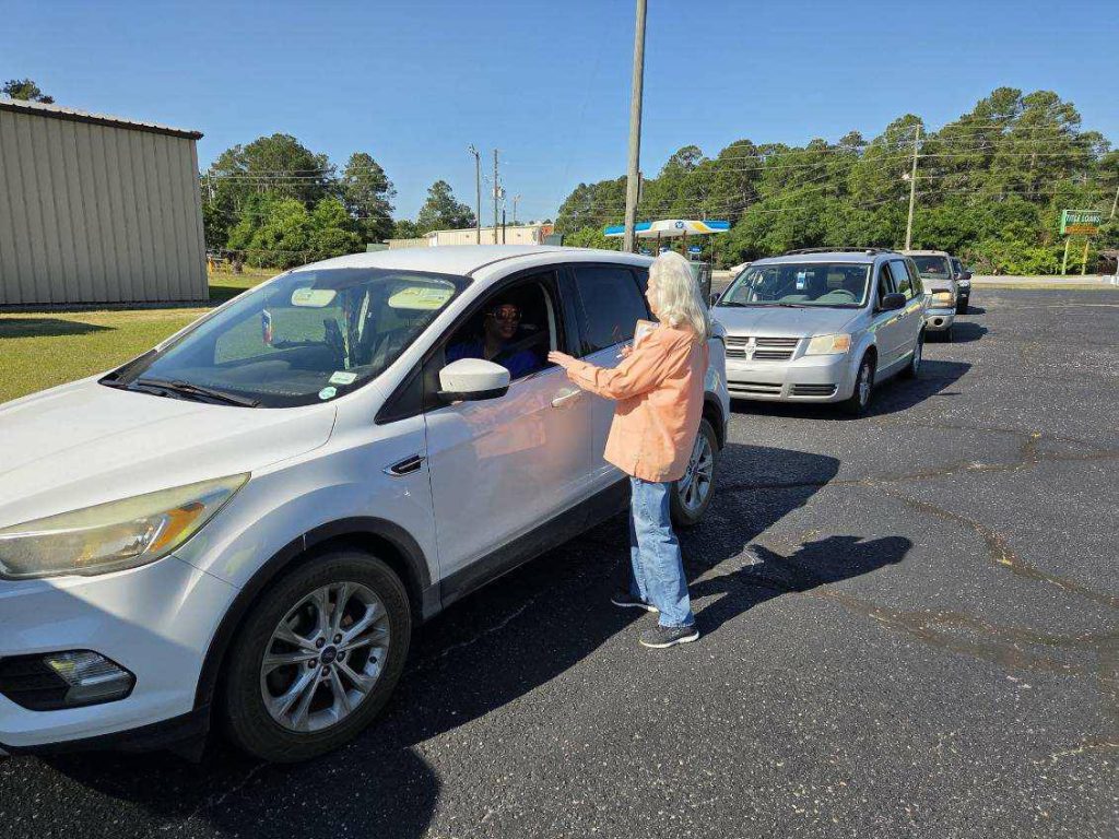 A woman hands something to a driver in a white SUV, with other cars lined up in a sunny parking lot.
