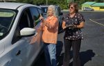 Two women stand next to a white car in a parking lot, one holding a clipboard, the other gesturing towards the driver.