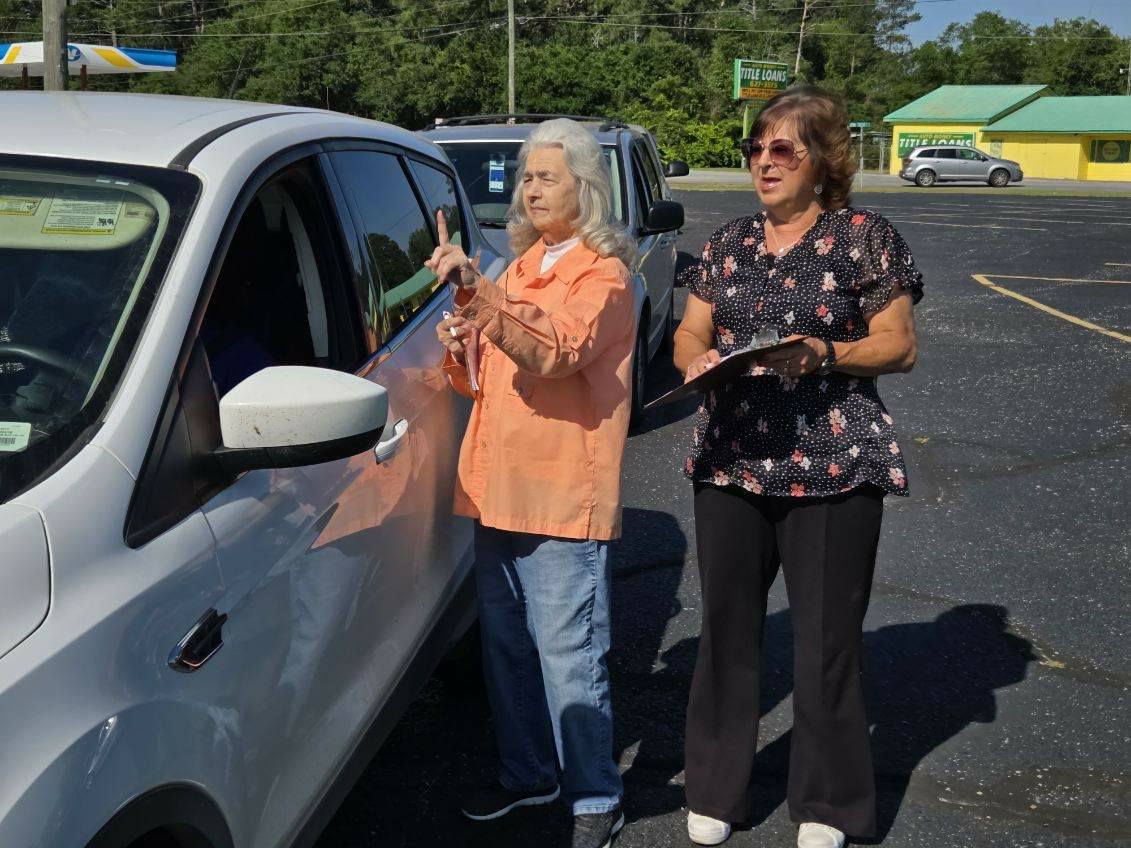 Two women stand next to a white car in a parking lot, one holding a clipboard, the other gesturing towards the driver.