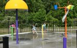 Children playing in a splash pad with colorful water features and fountains on a sunny day, surrounded by trees and a black fence.